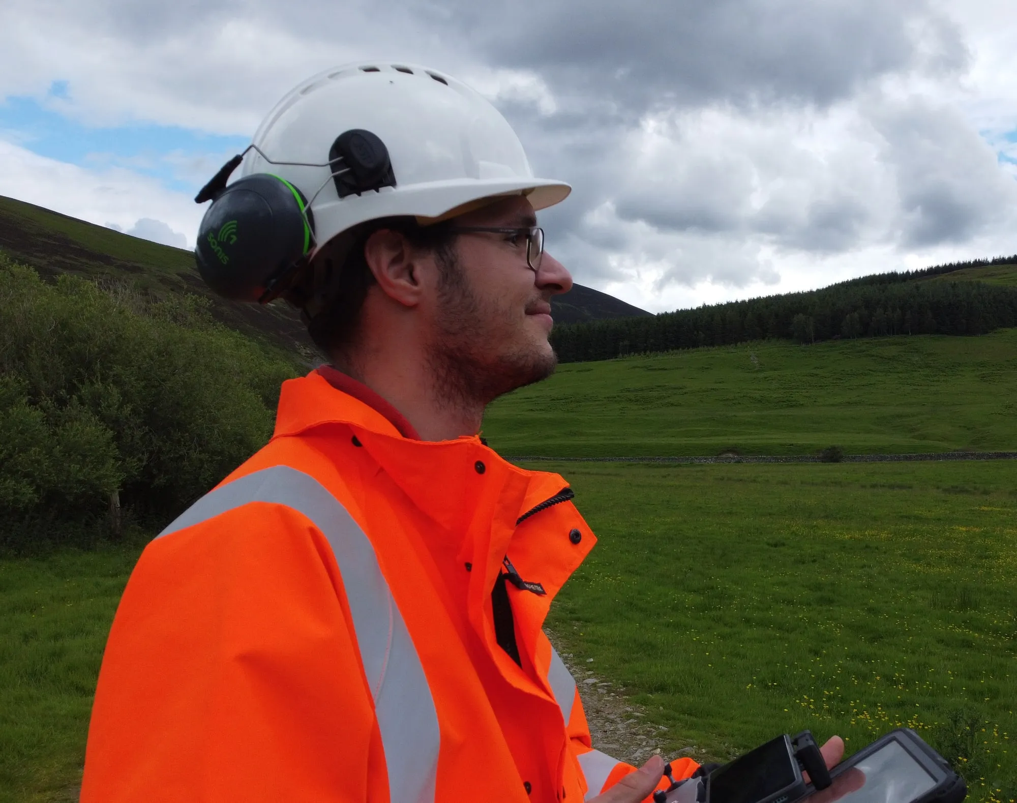 Leonluca Klein in a hard hat and hi-vis jacket, holding a tablet, with green hills and forest behind.